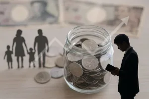 ガラス瓶に日本円の硬貨がぎっしり入った前景、右手前で財布を見つめるスーツ姿の若い独身男性、左奥には手をつなぐ4人家族のシルエット。背景にぼかした紙幣と上向きの矢印が重なり、独身税（子ども・子育て支援金）の負担増を象徴する写真